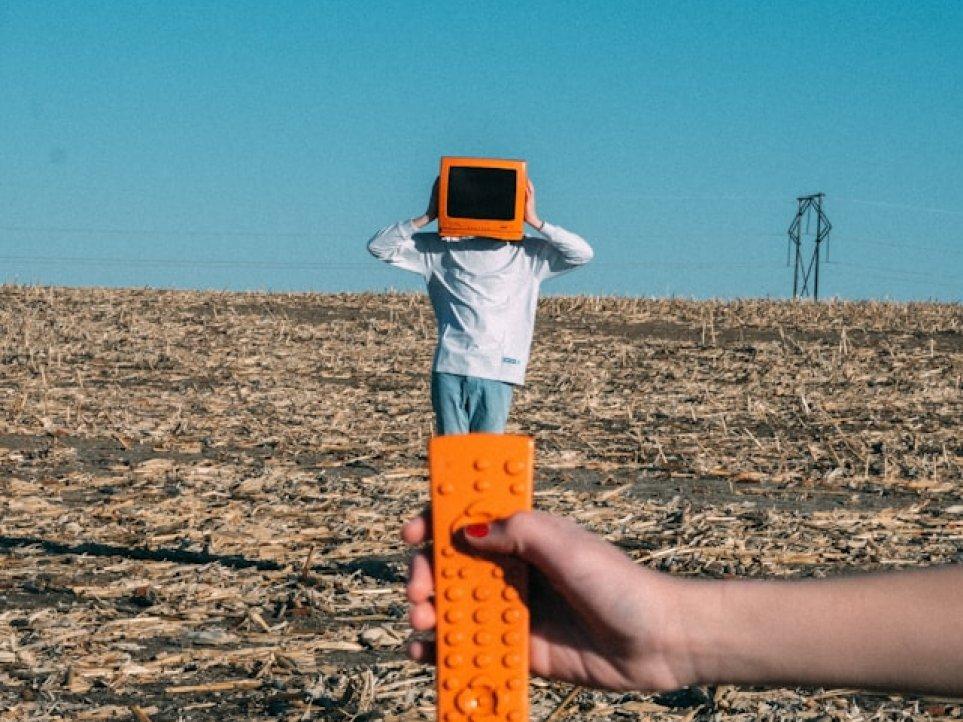 Eine Person in einem abgeernteten Feld mit orangefarbenem Monitor auf dem Kopf, während im Vordergrund eine Hand eine orange Fernbedienung auf sie richtet – ein Symbol für Kontrolle und Datensouveränität.