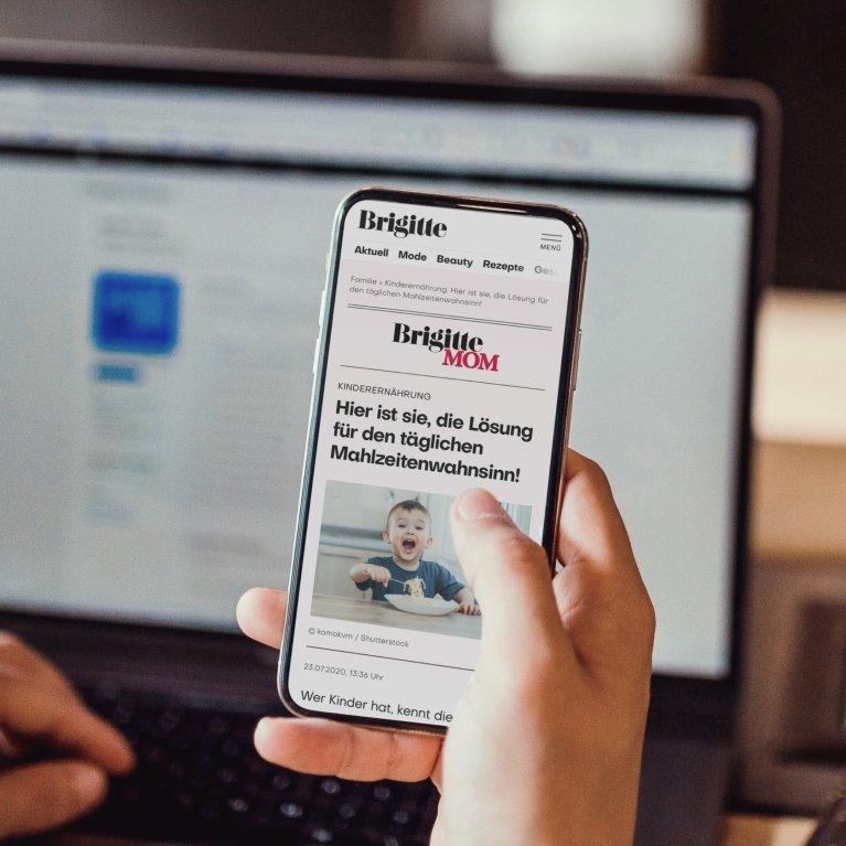 Eine Hand hält ein Smartphone, das die Brigitte Website zeigt. Im Hintergrund ist ein geöffneter Laptop zu sehen.
