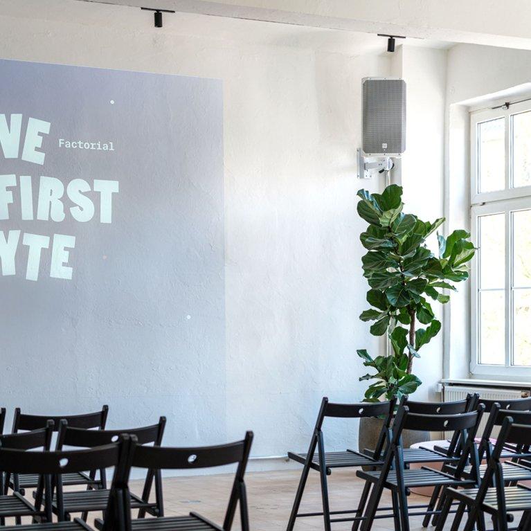 Modern event space at Factorial featuring a “Love at First Byte” projector screen, bright window fronts, indoor plants, and neatly arranged chairs ready for meetups or tech talks.