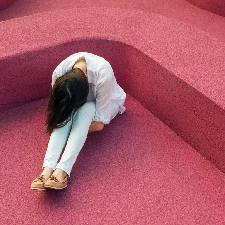 Woman sitting alone with head down on abstract pink structure – visual metaphor for emotional crisis, mental strain, and the need for compassionate digital support systems like TelefonSeelsorge.
