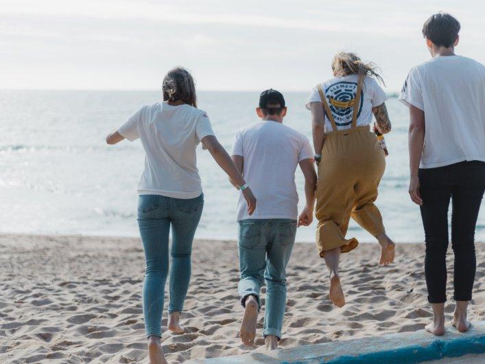 Vier Personen spazieren fröhlich an einem Sandstrand entlang, mit Blick auf das Meer und mit dem Rücken zur Kamera.